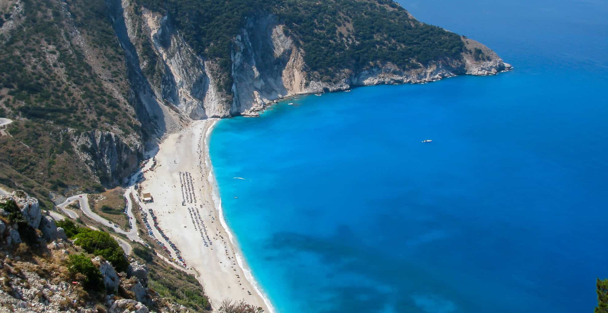 A high-angle view of Myrtos Beach, Kefalonia, with its brilliant turquoise water, long white pebble beach, and dramatic white cliffs.