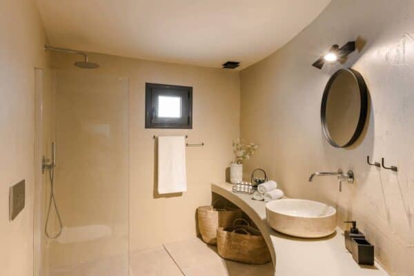 A Modern Bathroom With A Walk-in Glass Shower, A Concrete-style Sink Basin, A Round Wall Mirror, And Two Woven Baskets In Nomade Celes Suite Lixouri Kefalonia