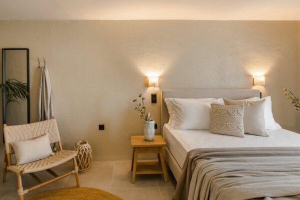 A modern, minimalist bedroom with a king-size bed, neutral-toned bedding, two wooden nightstands, and a stylish woven chair.