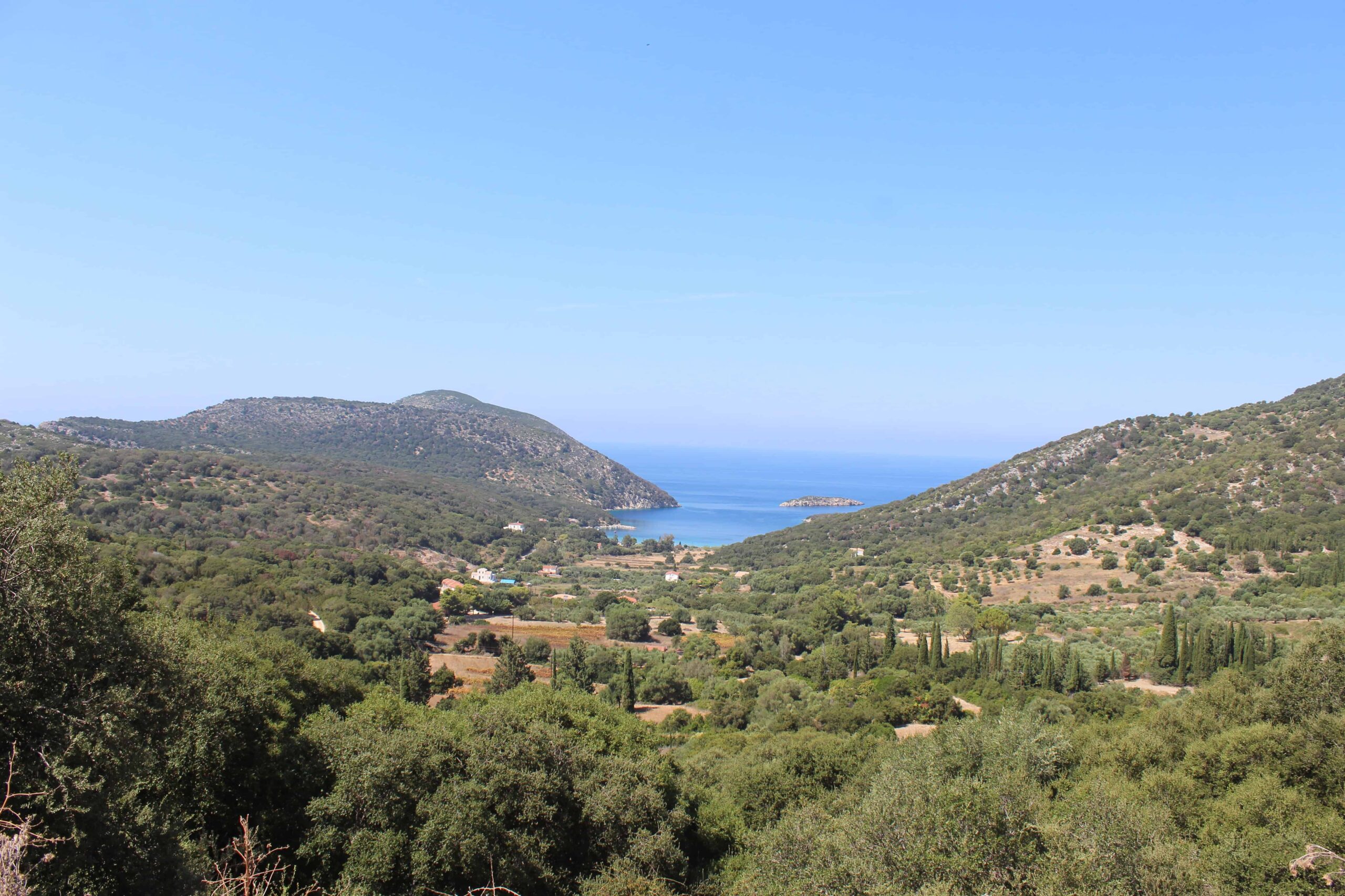 A panoramic view of Atheras beach from above, showing the bay nestled between two green, forested hills leading to the blue sea. Atheras Beach Kefalonia