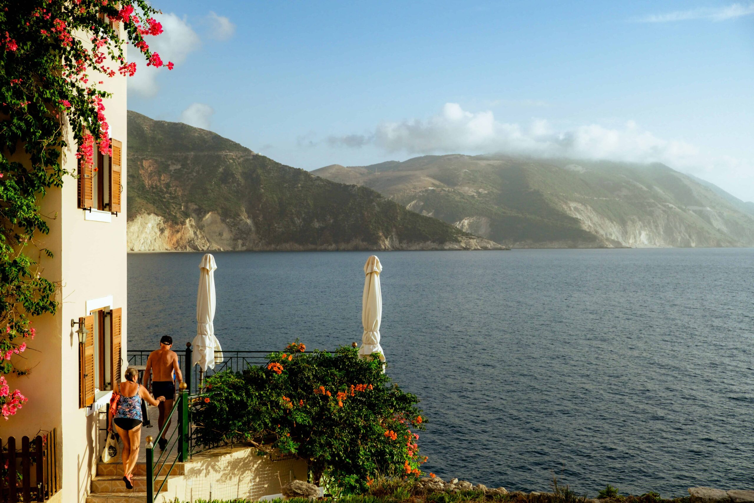 Two people walking down stairs from a building with pink flowers, towards a balcony overlooking the sea and distant mountains. christian-harb